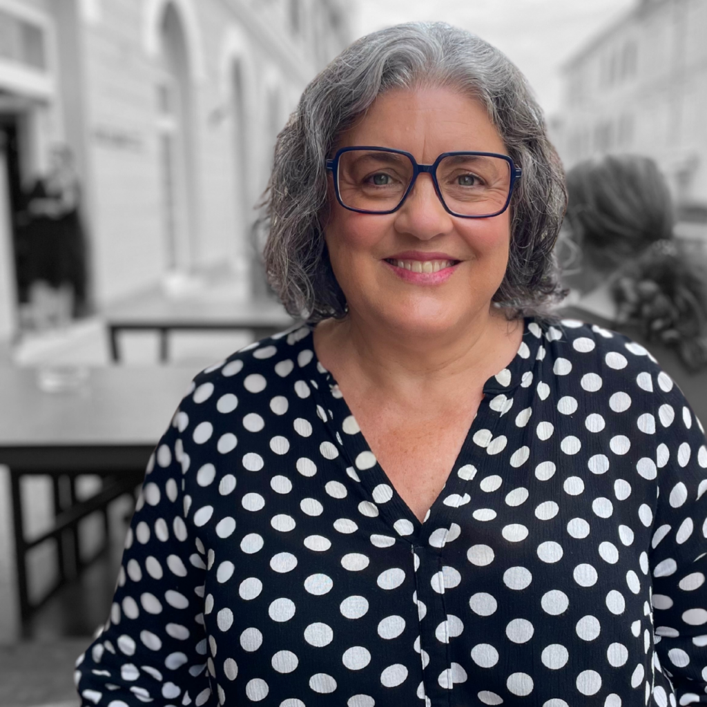 „Jutta Weiland mit grauen Locken und Polka-Dot-Bluse lächelt in die Kamera, farbig betont vor schwarz-weißem Straßenmotiv – Symbolbild für Persönlichkeit, Empathie und professionelle Begleitung.“